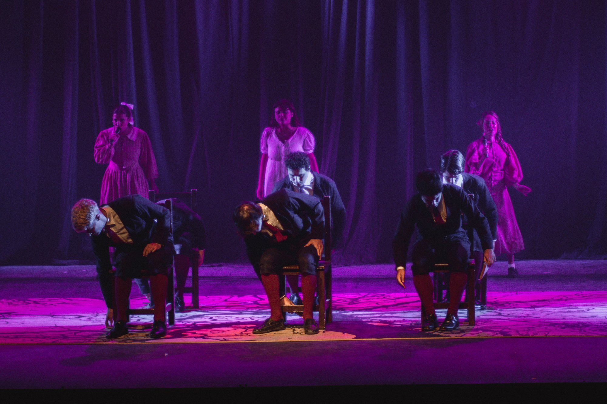 Escena con luz violeta: intérpretes sentados en sillas y otros de pie detrás.