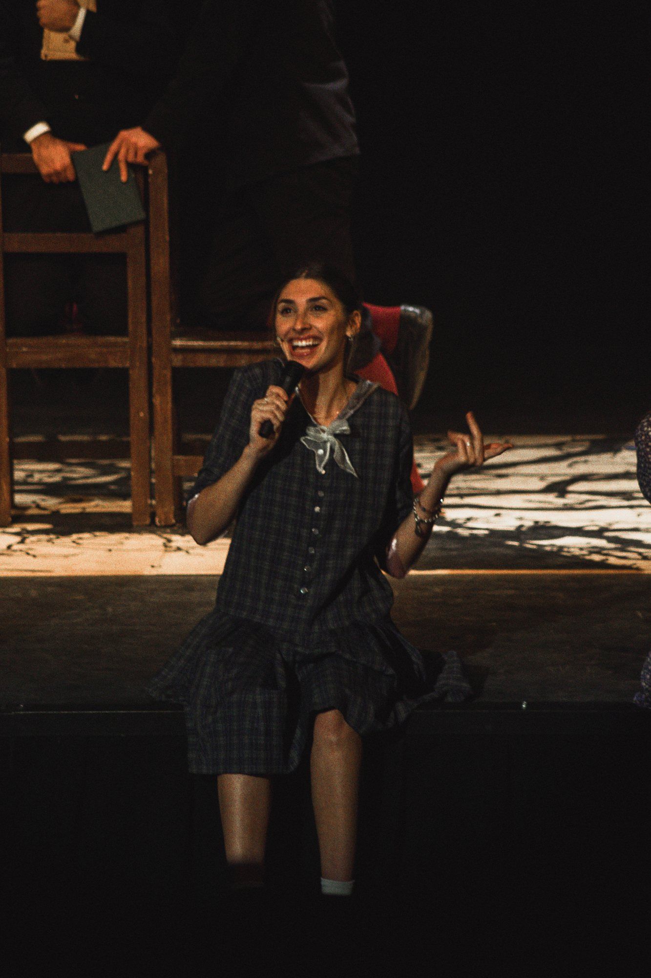 Una actriz canta con micrófono sentada en el borde del escenario.