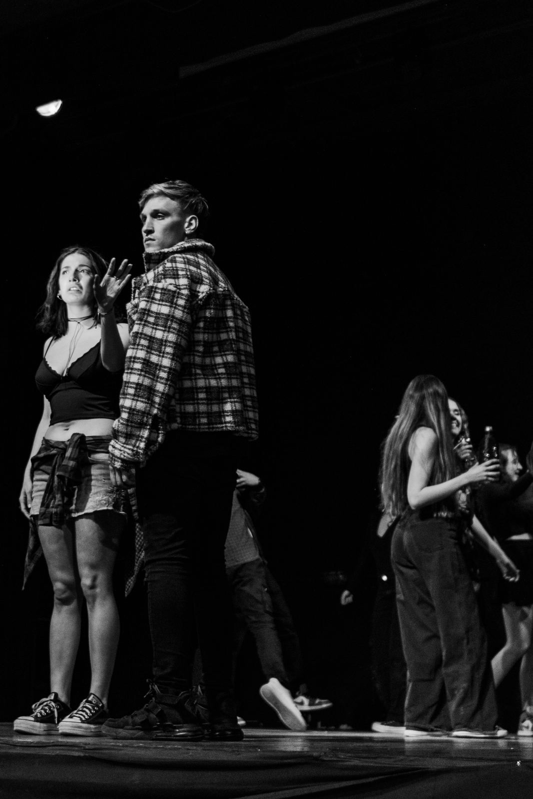 Escena de Dosis de Realidad en blanco y negro: una actriz y un actor de camisa a cuadros miran hacia fuera de escena; atrás, el ensamble.